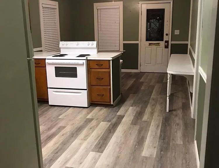 Small kitchen with stove installed in a narrow island, surrounded by light wood flooring. Walls are olive green with white trim and a door.