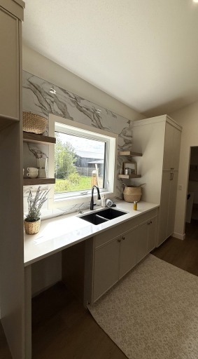 Bright, modern kitchen with white countertops and cabinets, a large window above a black sink, and decorative shelves holding wicker baskets and plants.