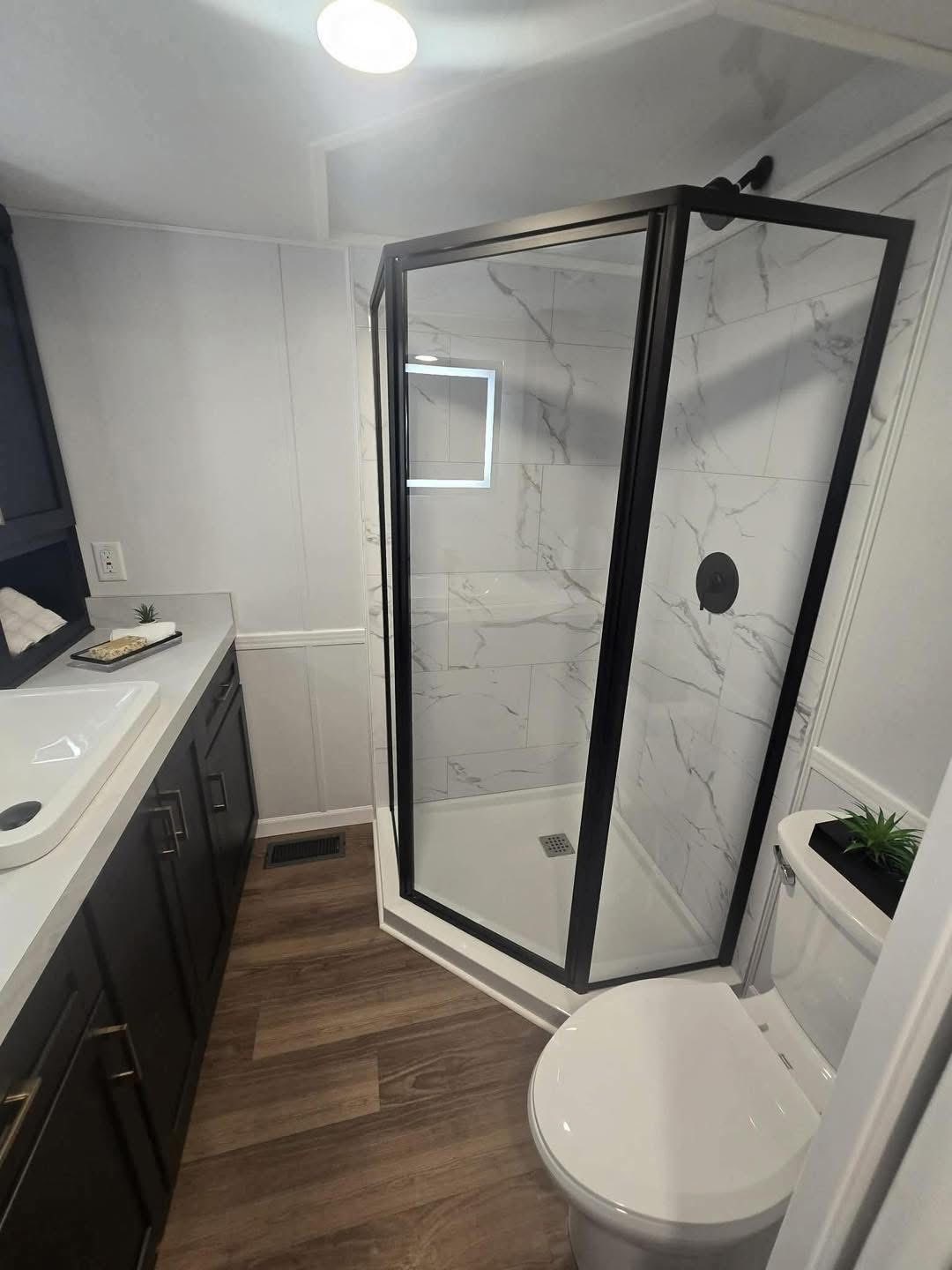 Modern bathroom with wood floor, glass shower with black frame and marble tiles. Dark vanity with white sink on left, white toilet with plant on right.