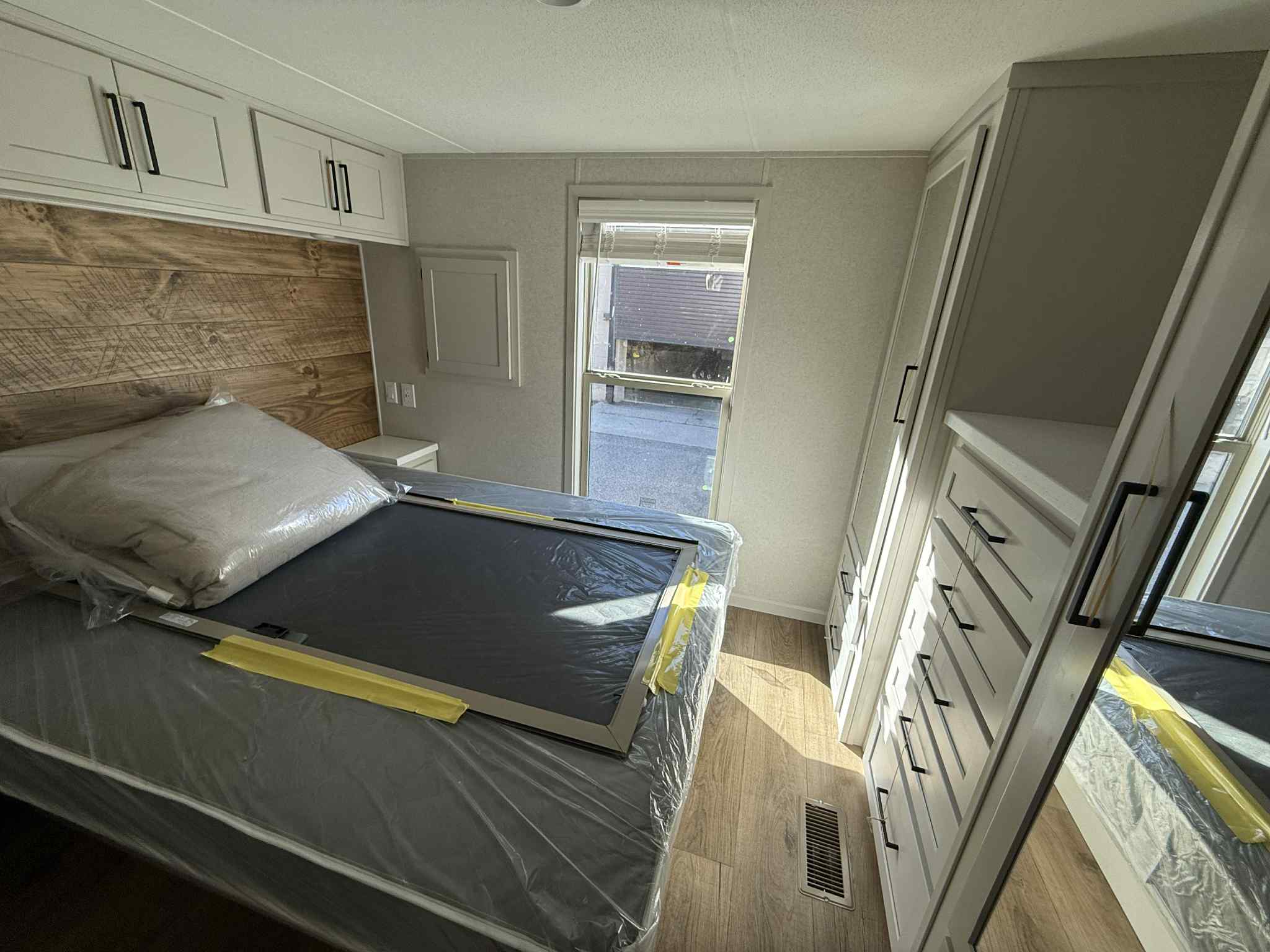 A compact bedroom with a neatly made bed, featuring wood accents and storage above. Large window on the right, bright natural light, minimal decor.