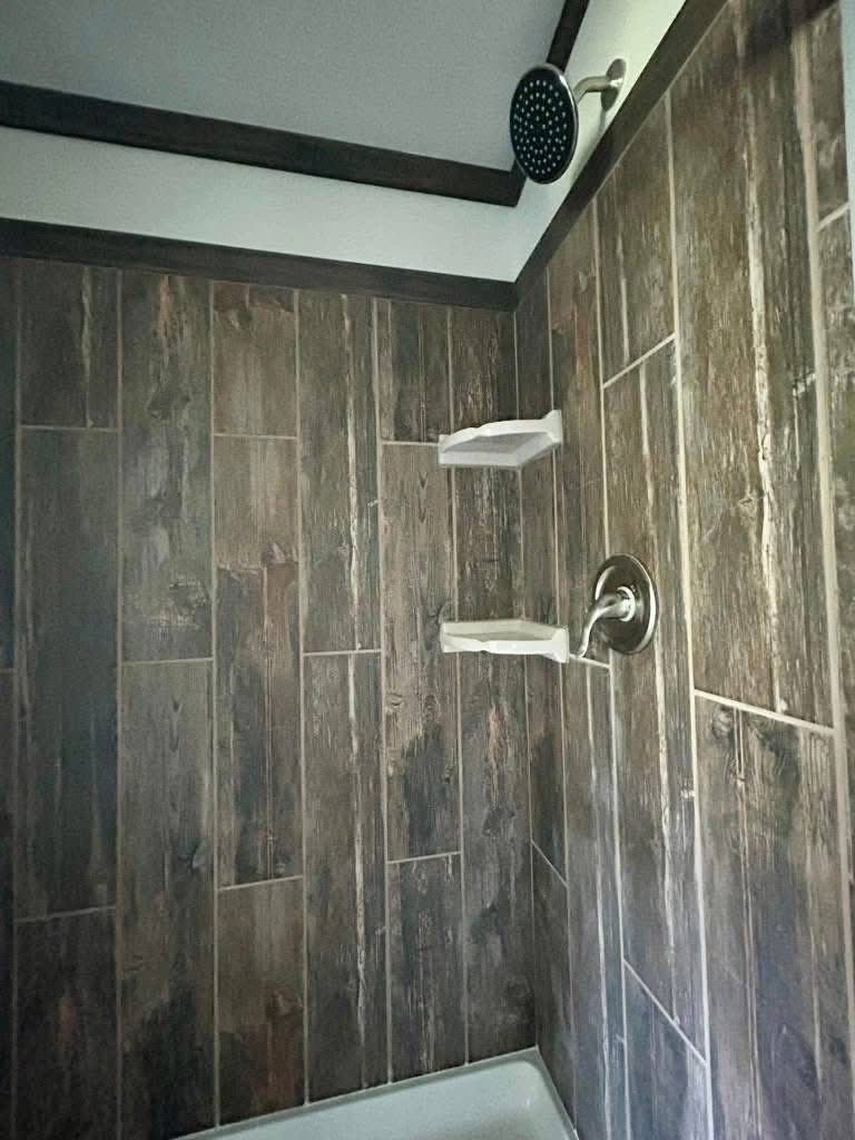Shower with rustic wood-patterned tiles, silver showerhead, and faucet. Two white corner shelves provide a modern and minimalistic ambiance.
