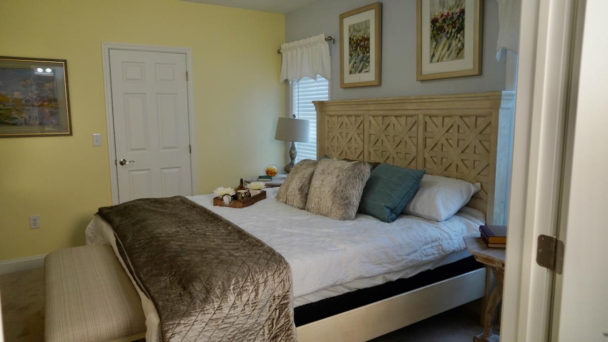 A cozy bedroom with a large bed, ornate wooden headboard, fluffy pillows, and a soft blanket. Warm yellow and beige walls enhance the inviting ambiance.