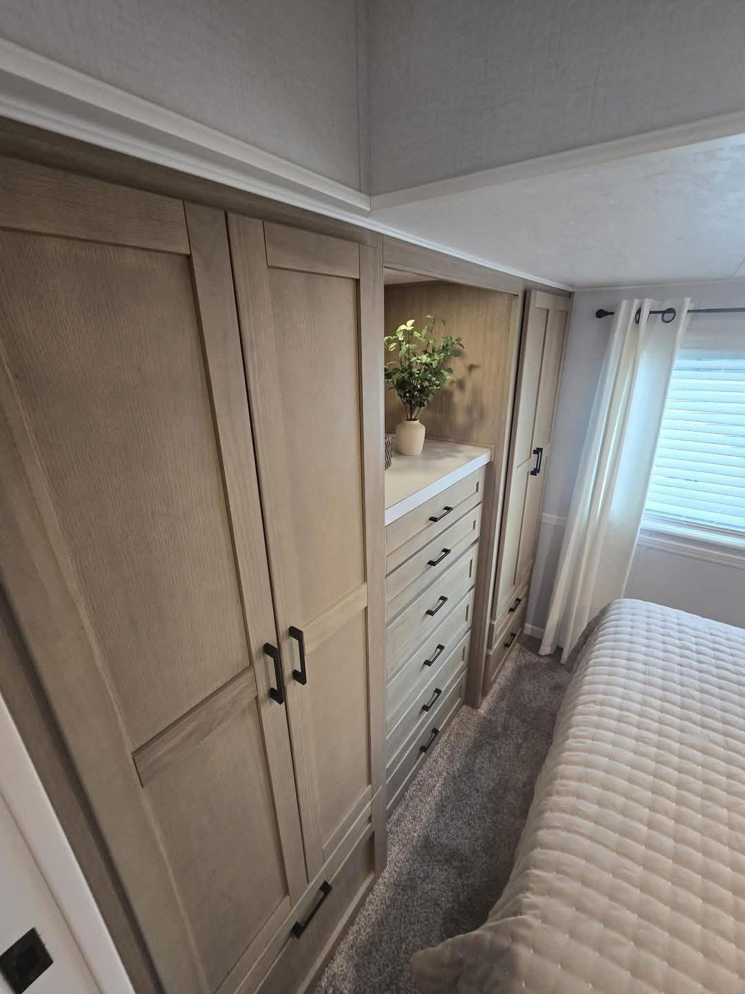 Bright bedroom with light wood cabinets and chest of drawers. A leafy plant decorates the top. Soft curtains and a textured bedspread add warmth.