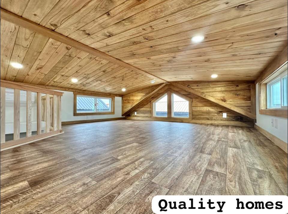 Spacious wooden loft with warm, rustic tones, features sloped ceiling, recessed lighting, and large triangular windows. Text reads "Quality Homes."