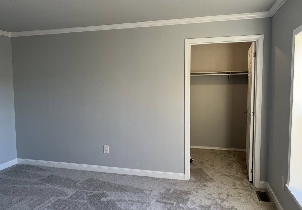 Empty room with light gray walls and carpet, featuring a white-trimmed doorway leading to a lit walk-in closet, evoking a simple, clean ambiance.