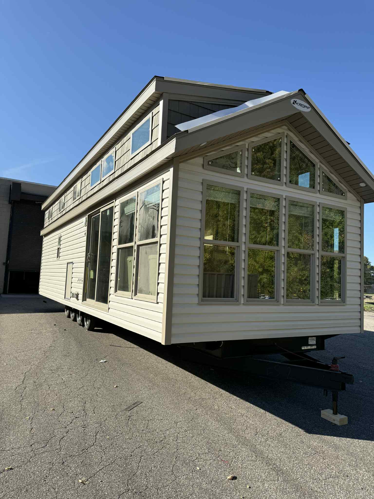 A mobile tiny home on wheels sits on an asphalt surface. It features a beige exterior, large windows, and is bathed in sunlight, suggesting mobility and freedom.