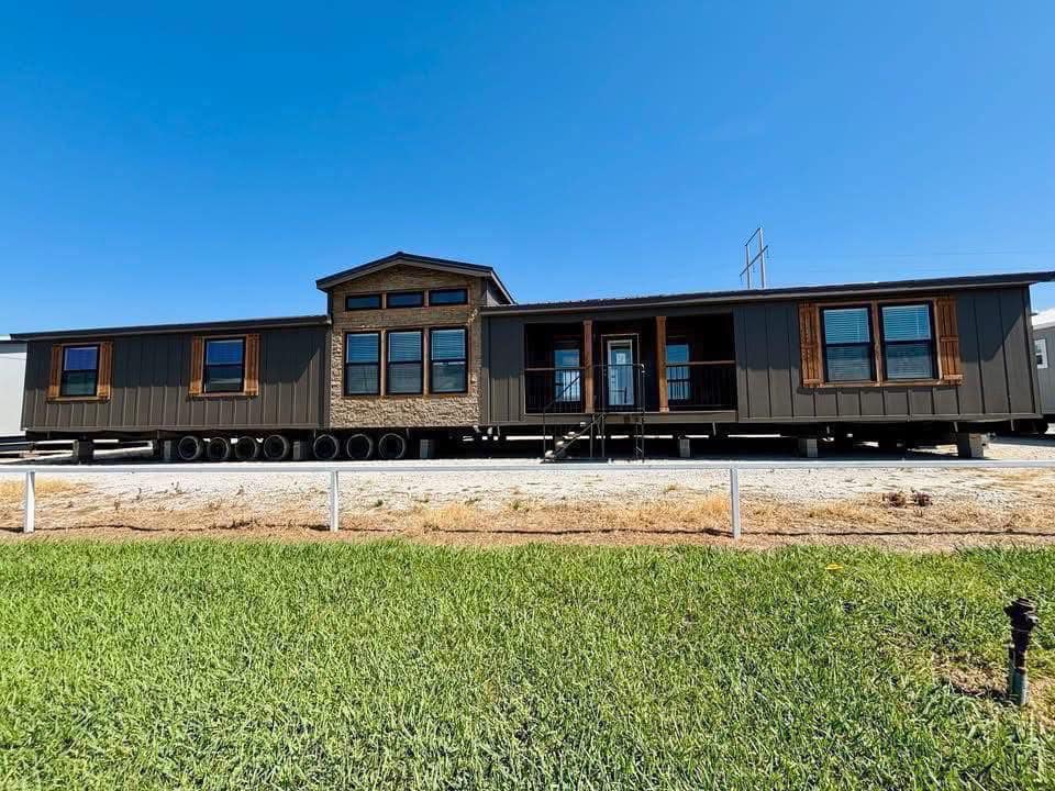 Brown manufactured home with a gabled roof and large windows, elevated on wheels. It is set against a clear blue sky, with grass in the foreground.