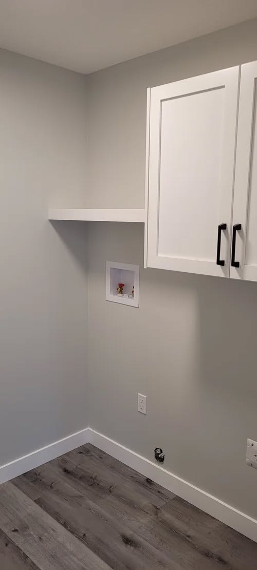 A minimalist laundry room corner with gray walls, a white cabinet, and a wooden floor. The space is empty, creating a clean, modern feel.