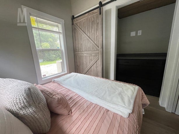 Bedroom with a pink bedspread and cozy pillows, facing a wooden sliding barn door. A window with a view of greenery brightens the serene space.
