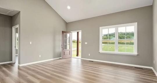 Spacious empty room with light wood flooring, gray walls, a large window, and a door opening to a scenic green view, conveying brightness and tranquility.