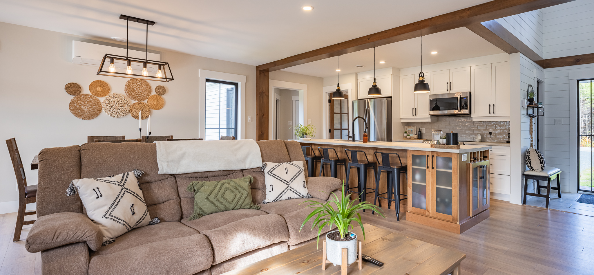 Modern open-concept living room and kitchen with natural tones. Features a brown sofa, stylish pendant lights, wooden beams, and a potted plant on the table.