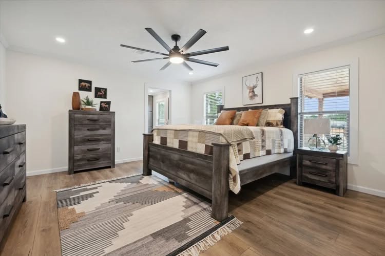 A modern bedroom with wooden flooring features a large bed, brown and beige quilt, rustic furniture, geometric rug, and deer artwork. The room is well-lit with a ceiling fan above, creating a cozy and inviting atmosphere.