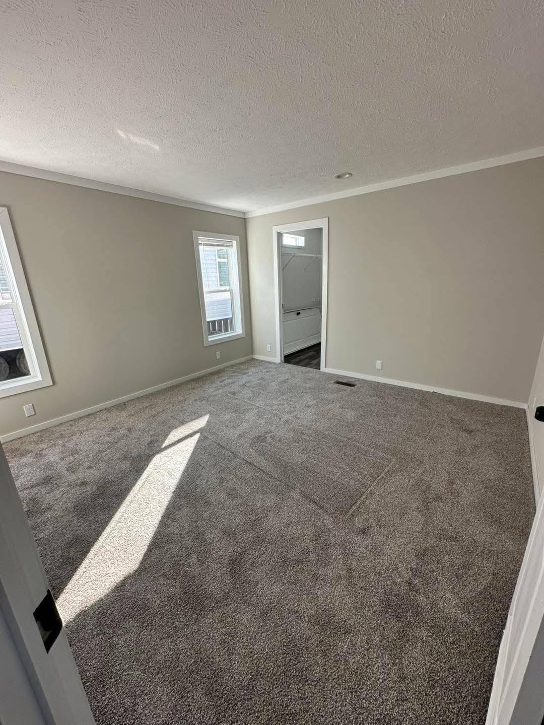 Empty room with beige walls, two windows, and gray carpet. Sunlight filters through, creating a warm ambiance. An open doorway leads to a bathroom.