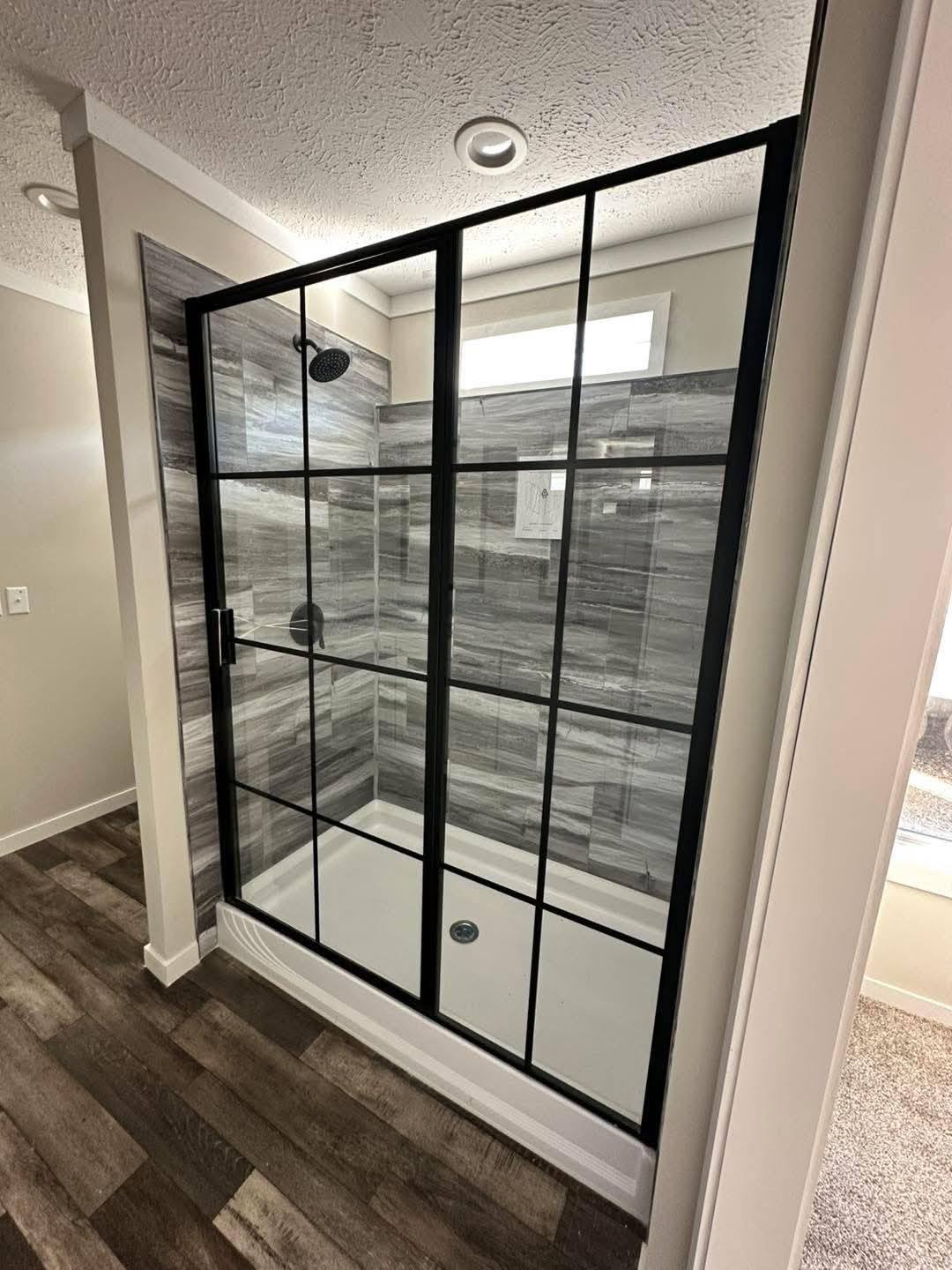Modern shower with grey marble tiles, black-framed glass door, and rainfall showerhead. Minimalist and clean design with wood-like flooring.