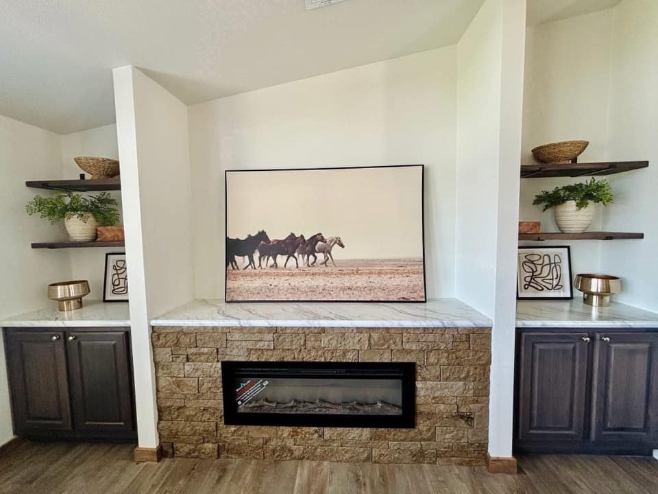 Cozy living room with a stone fireplace, flanked by dark wood cabinets. Above hangs a serene photo of horses on a plain. Shelves display plants and decor.