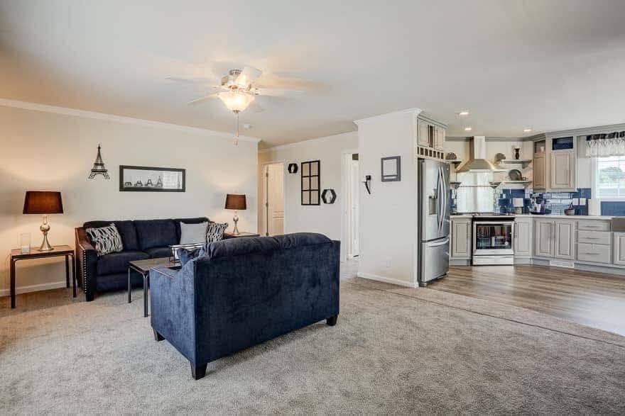 Spacious living room with navy sofas, wood flooring, and a ceiling fan. Open kitchen with stainless steel appliances and white cabinets. Cozy ambiance.