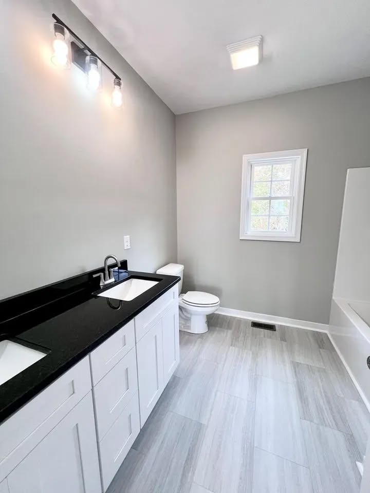 Bright bathroom with white cabinets and a black countertop under three-bulb lighting. Features a toilet, window, and bathtub. Modern and clean design.