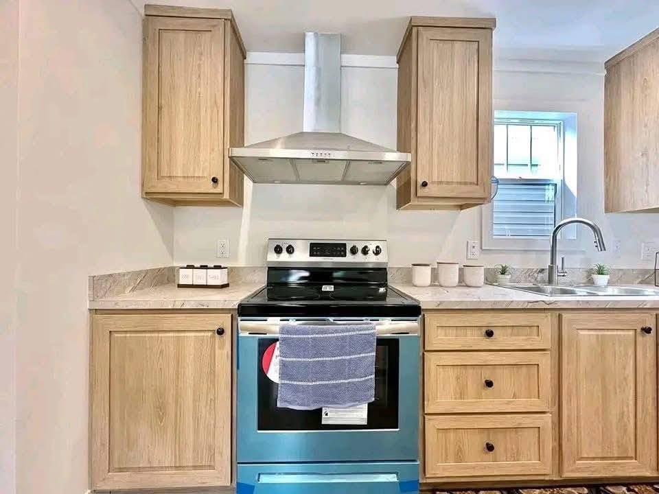 Modern kitchen with light wood cabinets and stainless steel appliances. An oven with a blue protective film and a towel hangs below a range hood. Bright, airy atmosphere.