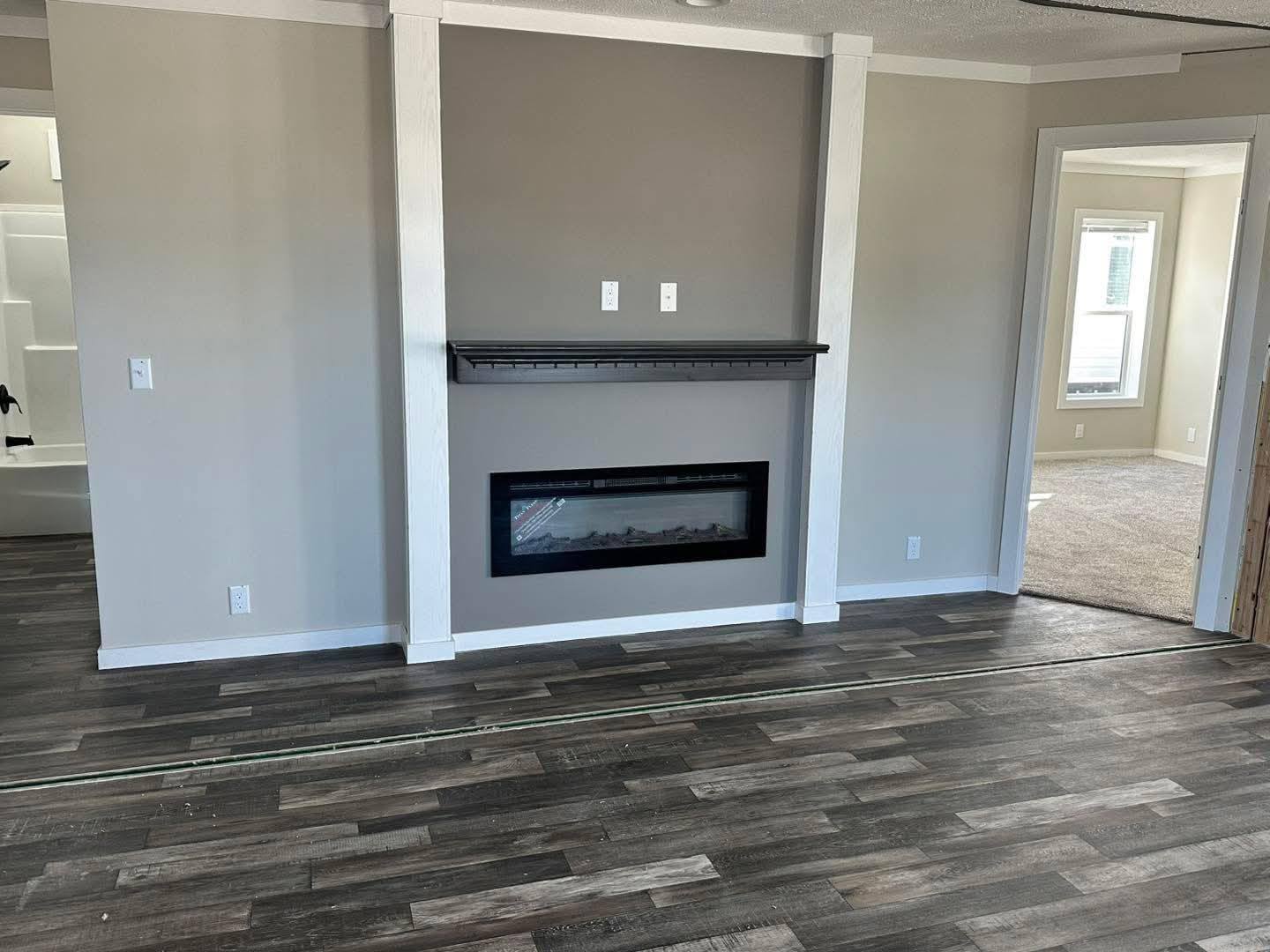 Modern living room with gray walls, dark wood floors, and a sleek electric fireplace. Doorway leads to a bright, carpeted room with large windows.
