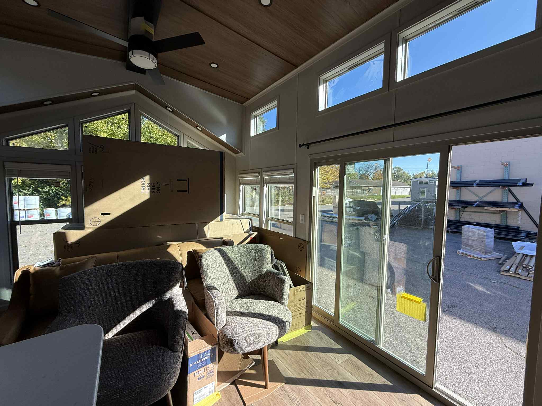Bright room with large windows and sliding glass doors, revealing an outside view. Multiple boxes and upholstered chairs fill the space. Ceiling fan above.