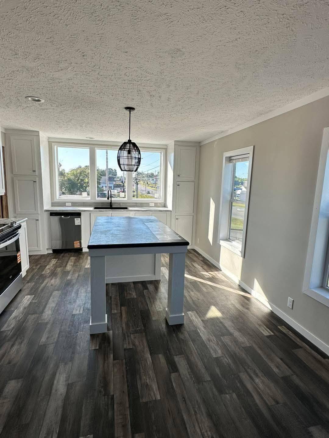 Bright kitchen with dark wood flooring, a central island with counter stools, and white cabinets. Large windows bring in sunlight, creating an airy feel.