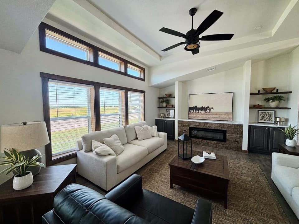 Stylish living room with beige sofas, a black armchair, and a wooden coffee table. Large windows illuminate a horse-themed painting above a stone fireplace, creating a cozy atmosphere.