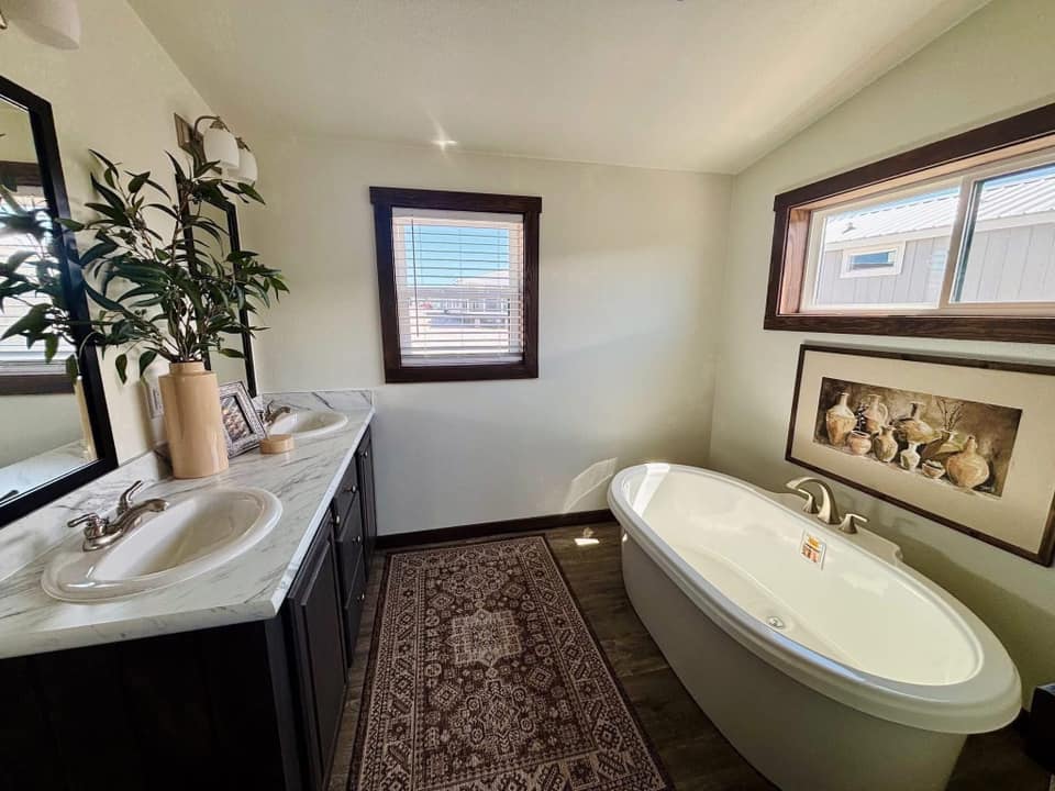 Bright bathroom with sunlight illuminating a white oval bathtub beneath a window. Features a double sink vanity with greenery, a decorative rug, and wall art.
