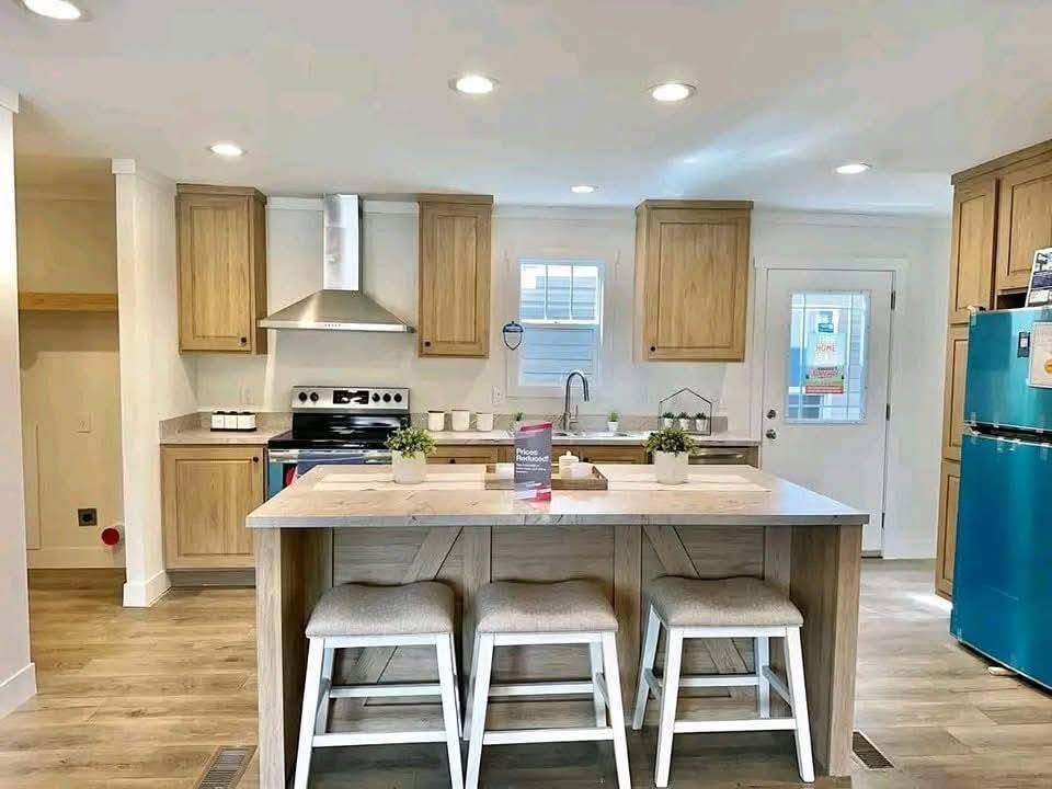 Bright kitchen with wooden cabinets, modern appliances, and a teal fridge. An island with three stools and potted plants creates a welcoming vibe.