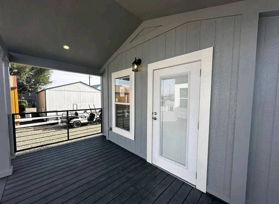 A gray wooden patio with a glass door and window framed in white. A small lantern light is above the door. Outside, a fenced yard with two sheds.