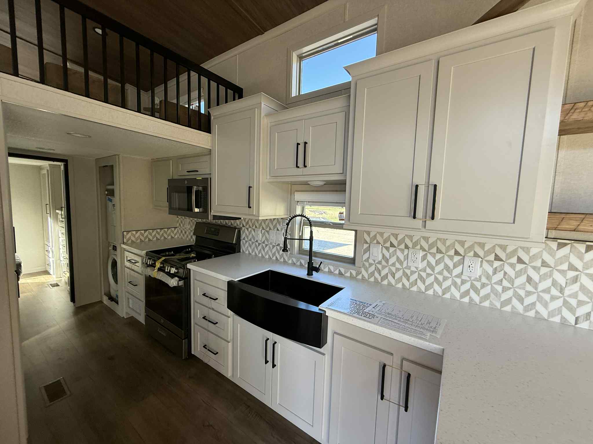 Modern kitchen in a tiny home with white cabinets, black sink, geometric backsplash, and stainless steel appliances. Upper loft visible above.