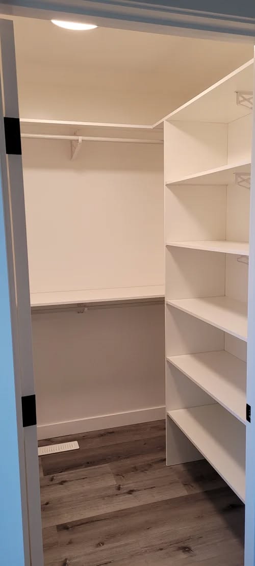 A spacious, walk-in closet with white shelves on the right and a clothes rod against the back wall. The wooden floor and bright lighting create an inviting ambiance.