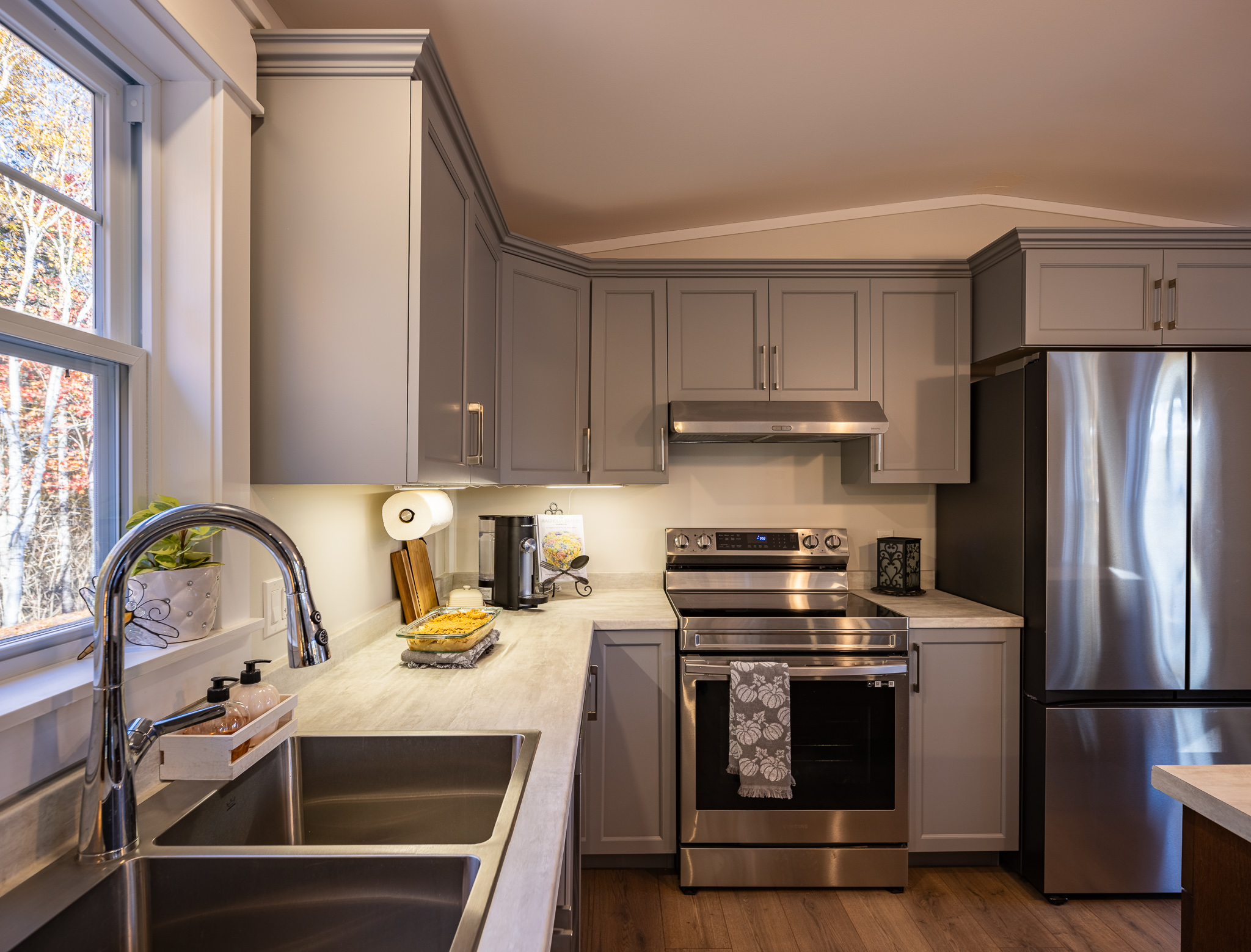 Modern kitchen with gray cabinets, stainless steel appliances, dual sinks, and warm lighting. A window gives a glimpse of fall foliage outside.
