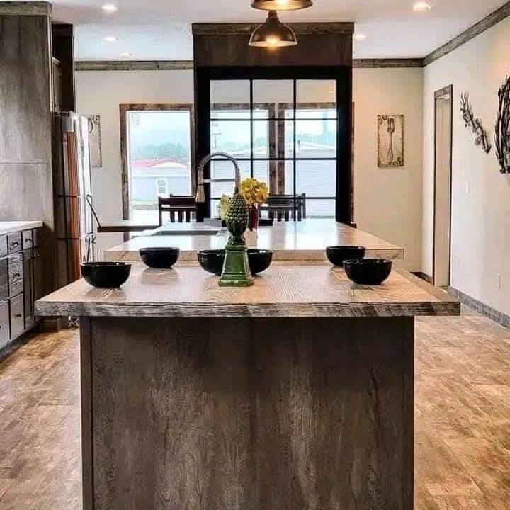 Modern kitchen with a rustic touch featuring a central island topped with black bowls and a vase of flowers. Large windows allow natural light in.