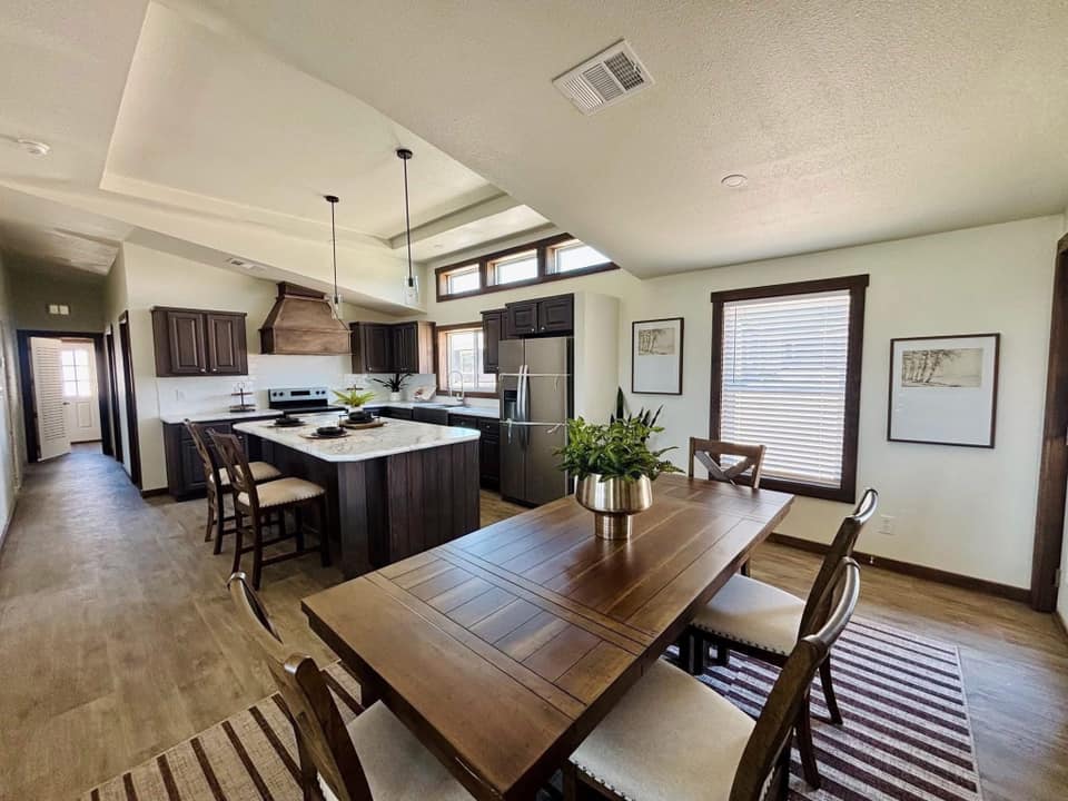 A modern kitchen and dining area with warm wooden tones. Features an island, hanging lights, potted plant, window blinds, wall art, and a striped rug.