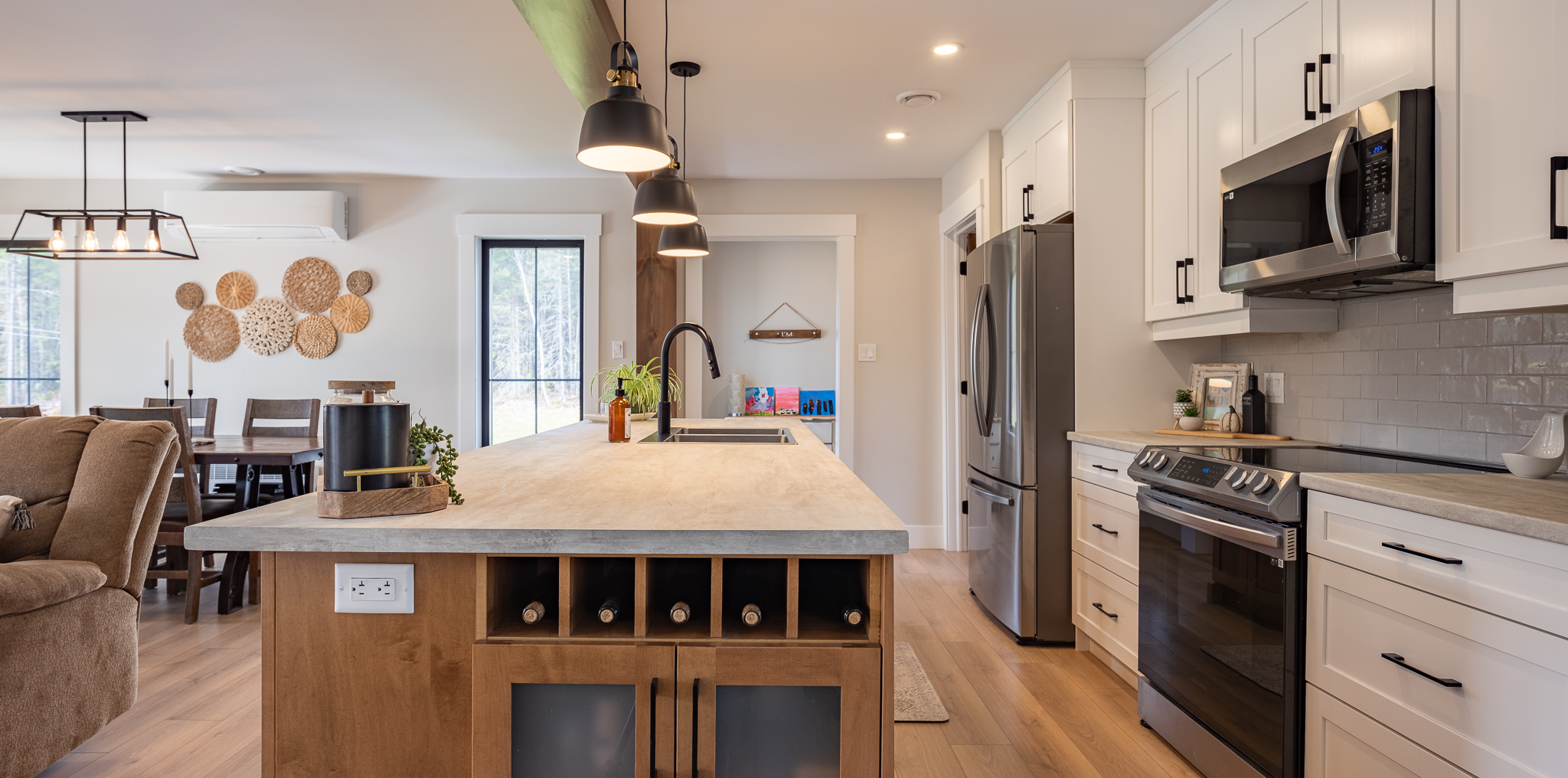 Modern kitchen with a central island featuring wine storage, pendant lights, and a sink. White cabinets, stainless steel appliances, and wooden accents create a warm, inviting atmosphere. Dining area visible in the background.