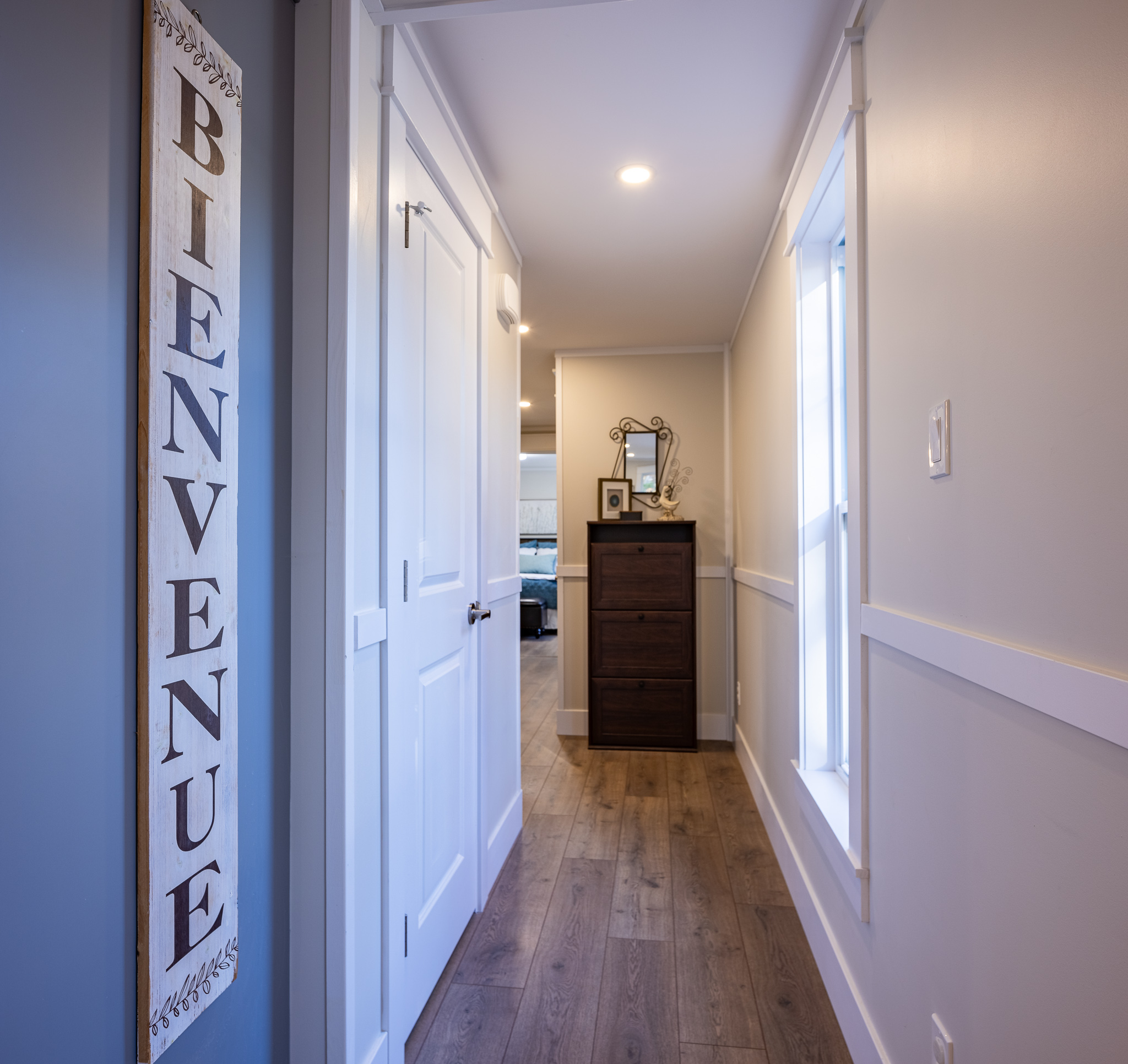 Narrow hallway with wooden floors and a "Bienvenue" sign on the wall. A chest of drawers with decor is at the end; bright and welcoming atmosphere.