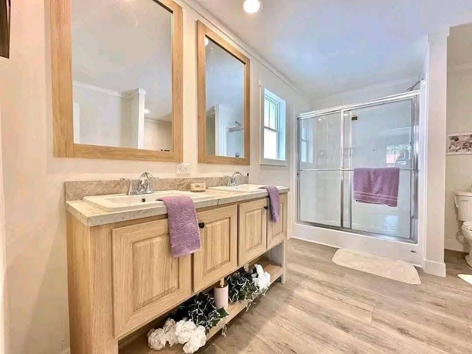 Bright, modern bathroom with dual sinks, wooden vanity, large mirrors, glass shower door, purple towels, and decorative white flowers on the shelf below.