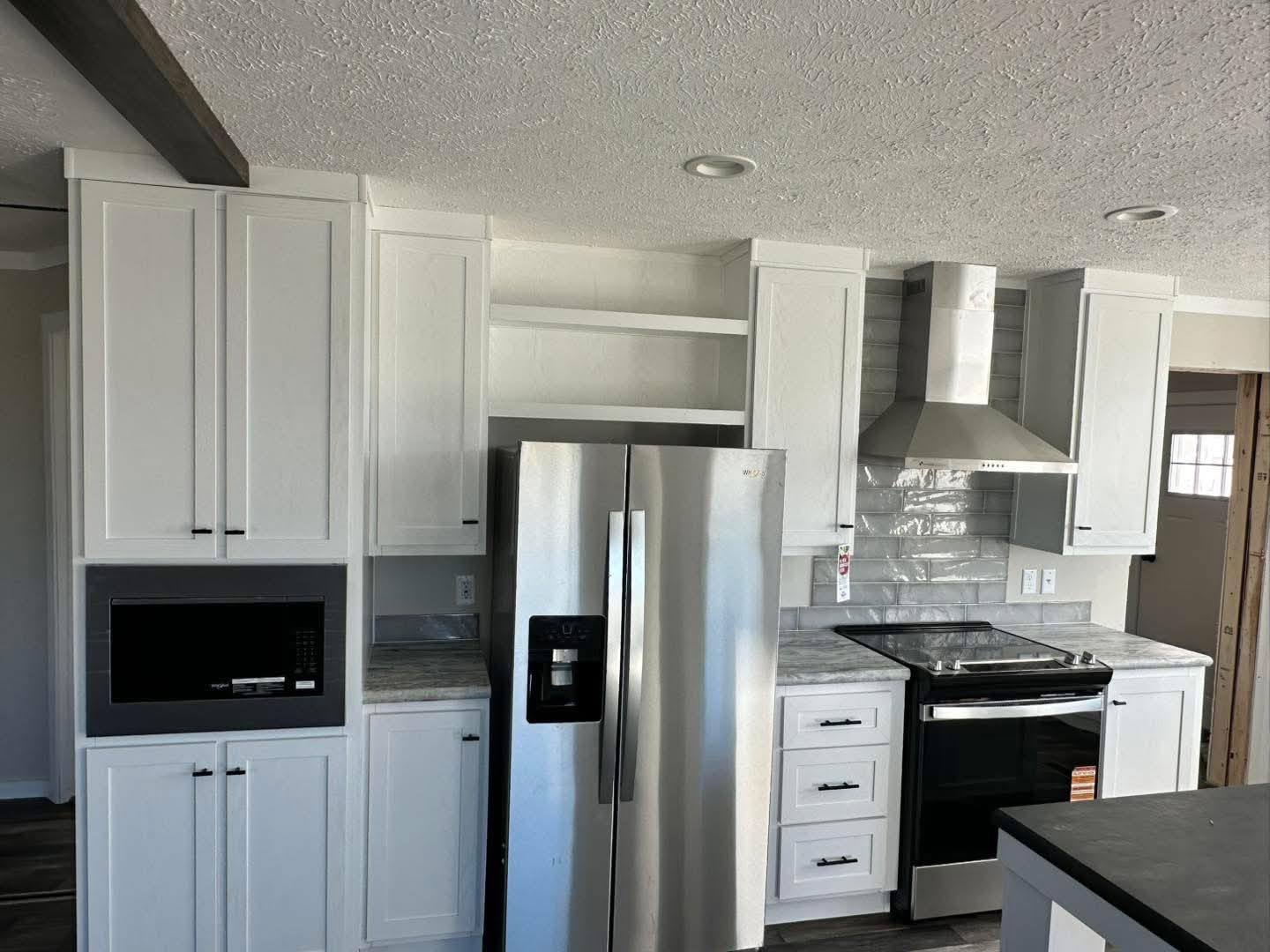 Modern kitchen with white cabinets, stainless steel appliances including a refrigerator, oven, and range hood. Gray backsplash tiles add contrast.