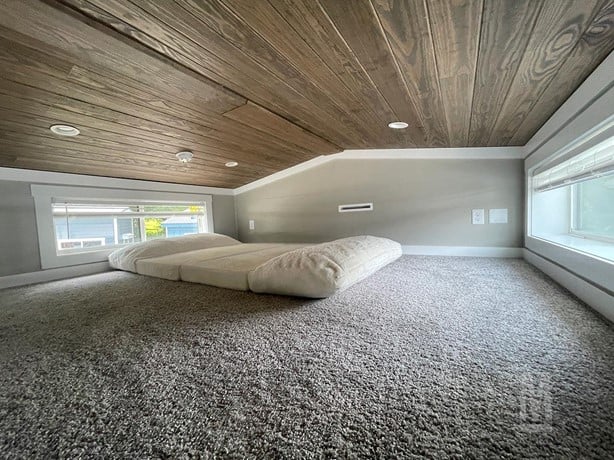 Cozy loft space with a sloped wooden ceiling and light gray carpet. A low mattress lies on the floor, flanked by two narrow windows. Calm and snug.