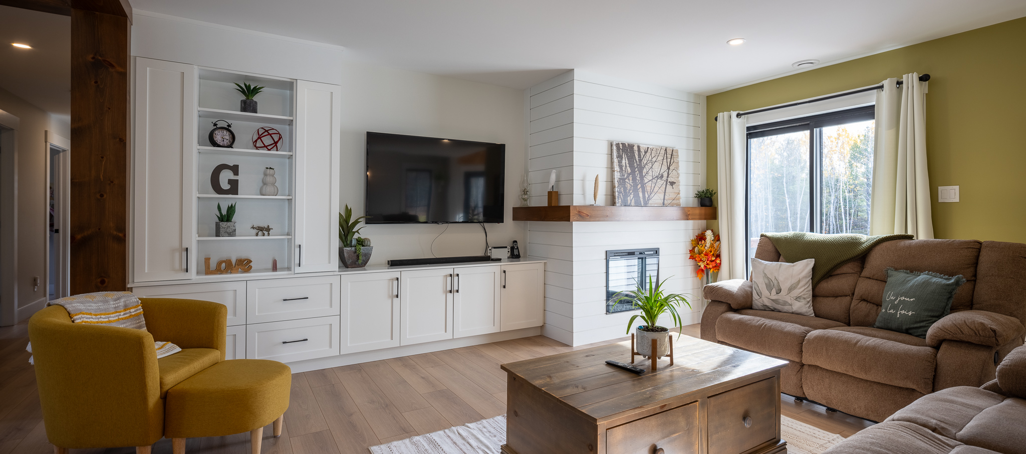 Modern living room with white walls, a mounted TV, built-in shelves, and a cozy fireplace. Features mustard chair, brown sofa, and large window.