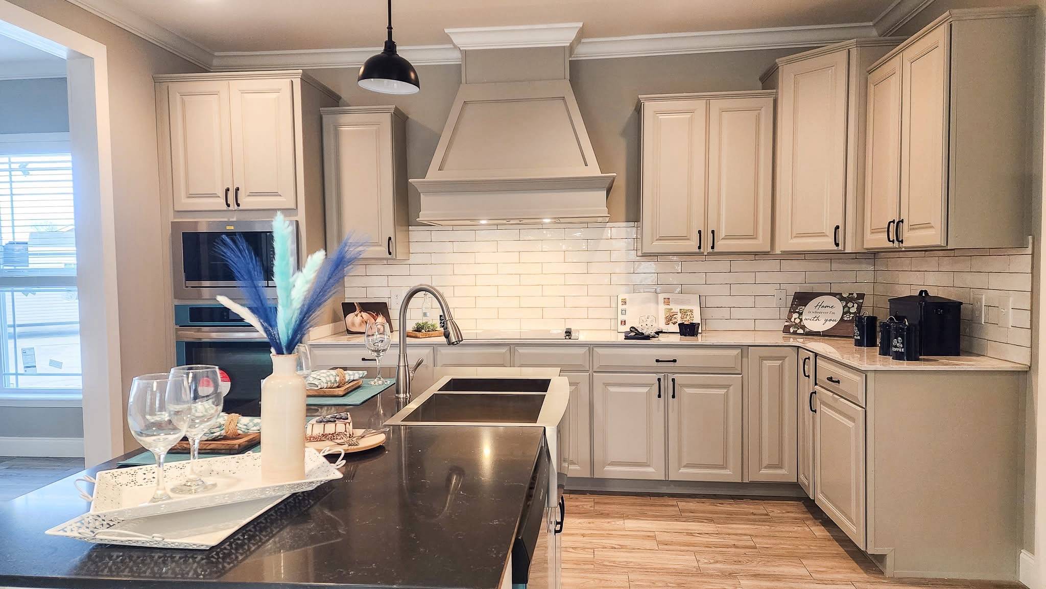 A modern kitchen with beige cabinets, white subway tile backsplash, and black countertop featuring decorative vase and wine glasses. Warm, inviting atmosphere.
