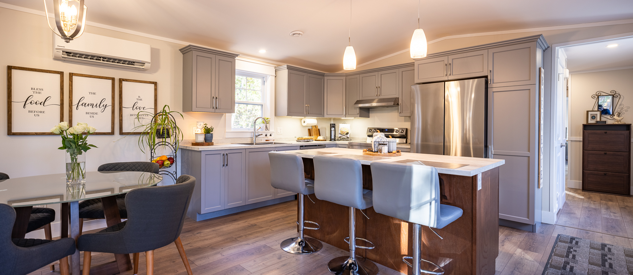 Modern kitchen with gray cabinets, stainless steel appliances, and a wooden island with three stools. The dining area has a round table with gray chairs and a flower vase. Warm lighting and decorative wall art create a cozy atmosphere.