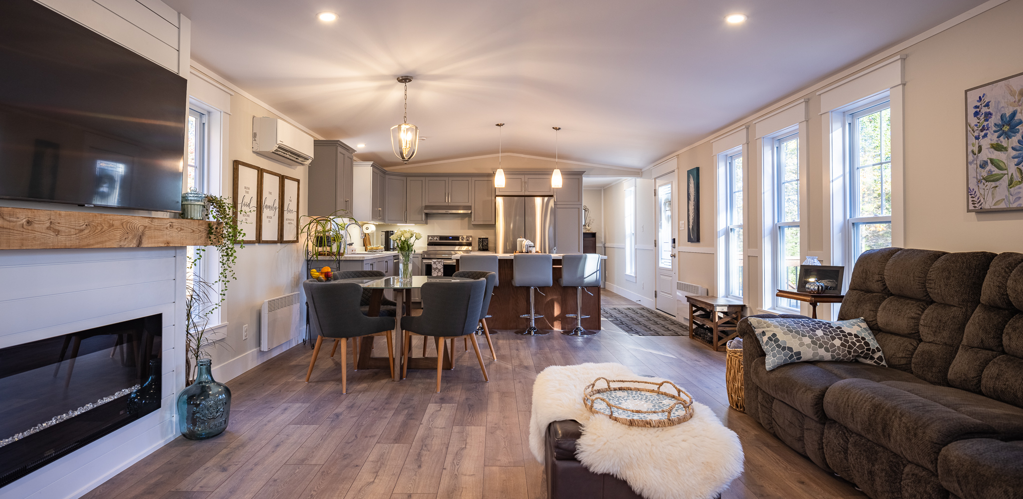 A cozy open-plan living room and kitchen with warm wood floors. Features include a plush sofa, a large TV, modern kitchen island with stools, and soft lighting.