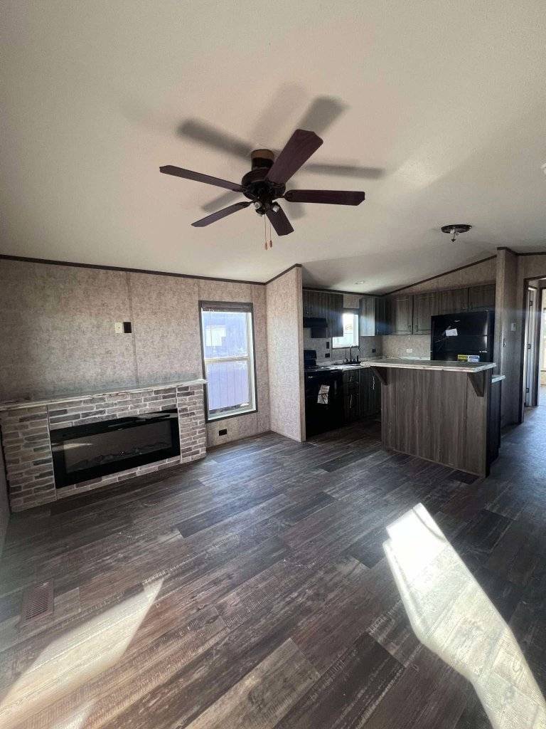 Spacious room with wood floor, ceiling fan, and fireplace on the left. Kitchen with dark cabinets and modern appliances on the right, creating a cozy atmosphere.