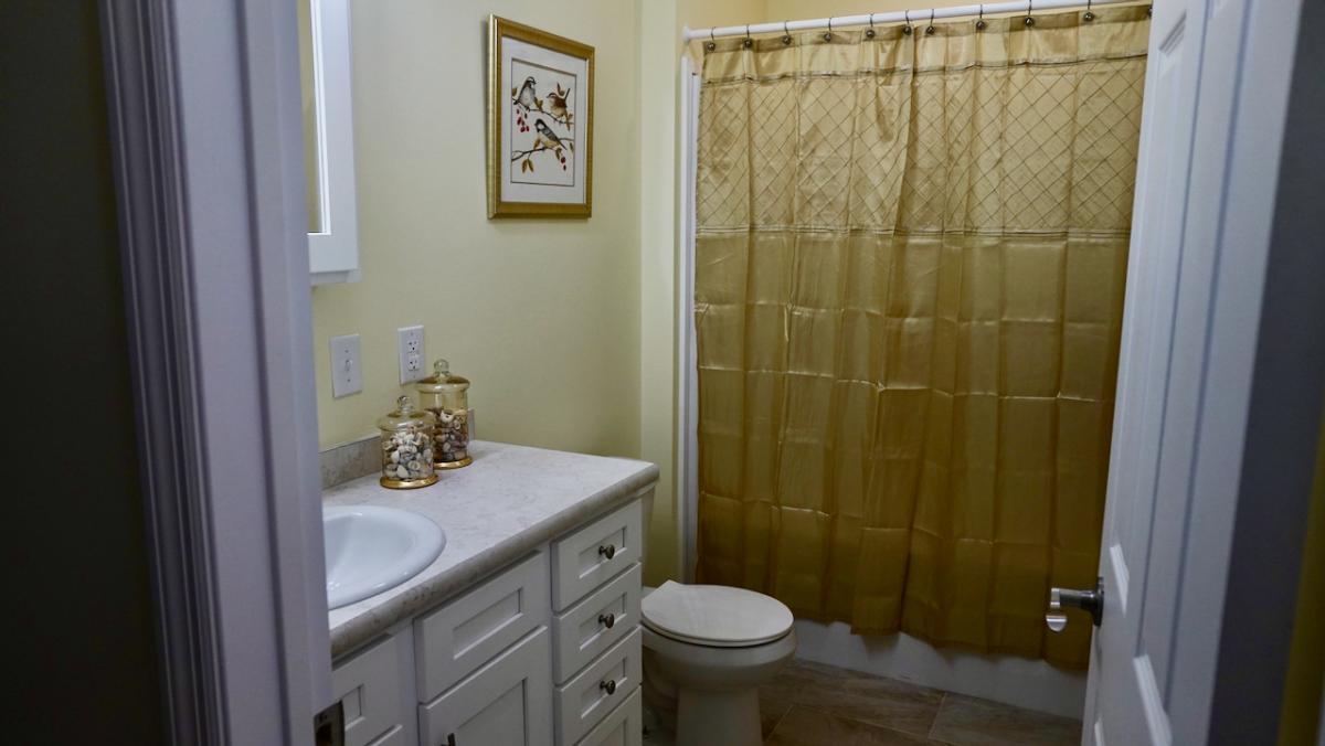 A small, well-lit bathroom with pale walls, featuring a white vanity and sink. A gold shower curtain and framed art add a touch of elegance.