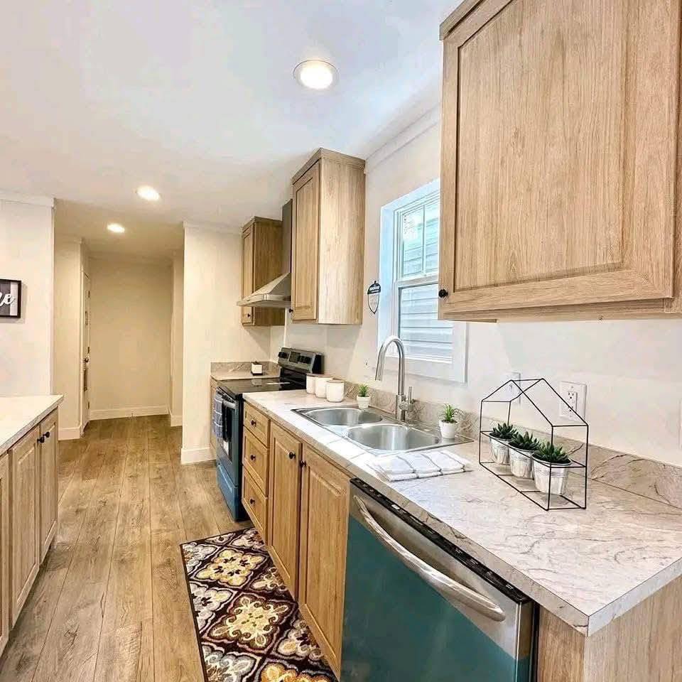 Modern kitchen with light wood cabinets, marble countertops, and stainless steel appliances. A colorful rug and small potted plants add warmth.
