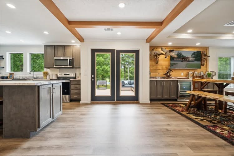 Spacious kitchen and dining area with wooden beams. Gray cabinets, stainless steel appliances, and large glass doors reveal a lush green exterior.