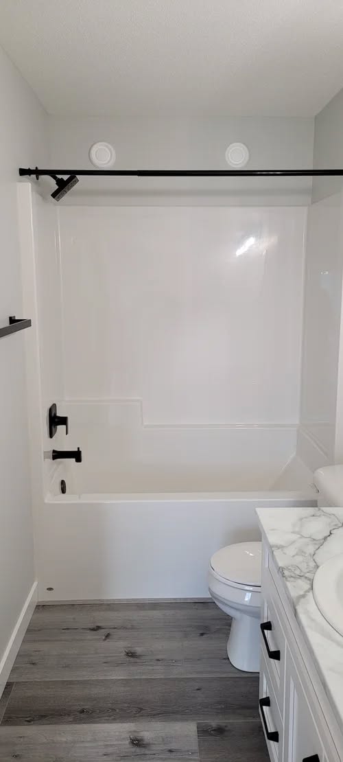 Modern bathroom with a white tub and shower featuring black fixtures. Grey wood floor, marble sink countertop, white walls, and a minimal aesthetic.