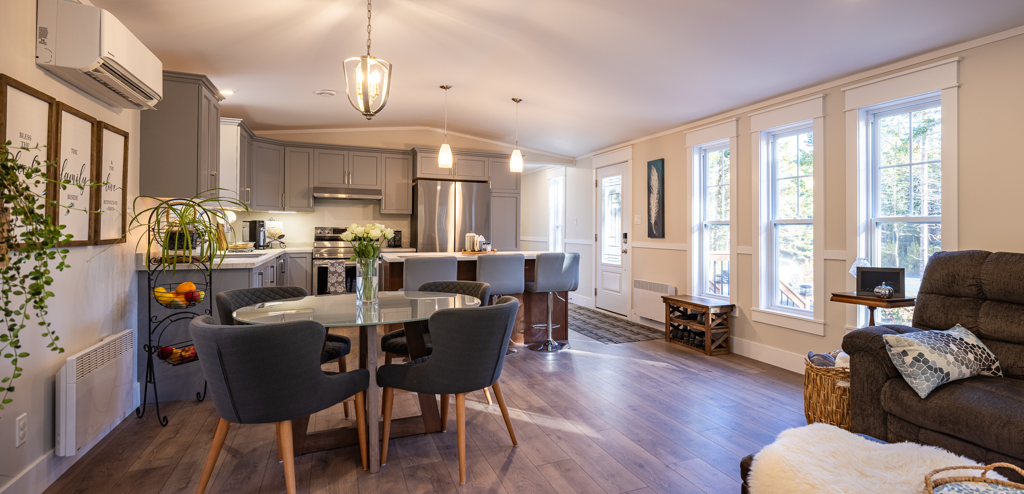 Modern open-plan kitchen and living area with gray cabinets, dining table, and cozy seating. Natural light from large windows, creating a warm, inviting atmosphere.