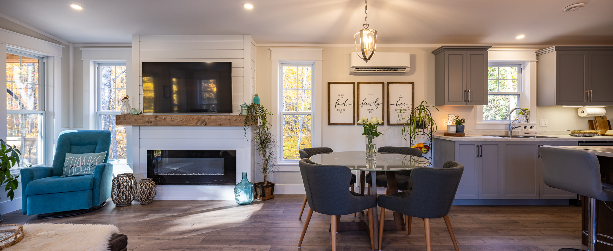 Stylish living area with a wall-mounted TV above a fireplace, teal armchair, and round dining table with flowers. Gray kitchen cabinets add elegance.